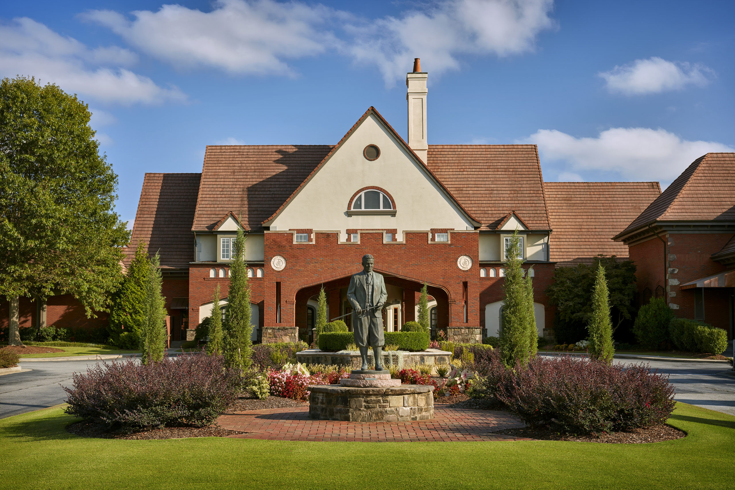 Red brick campus building with a steep tiled roof and central statue in a circular flower bed under a blue sky with scattered clouds.