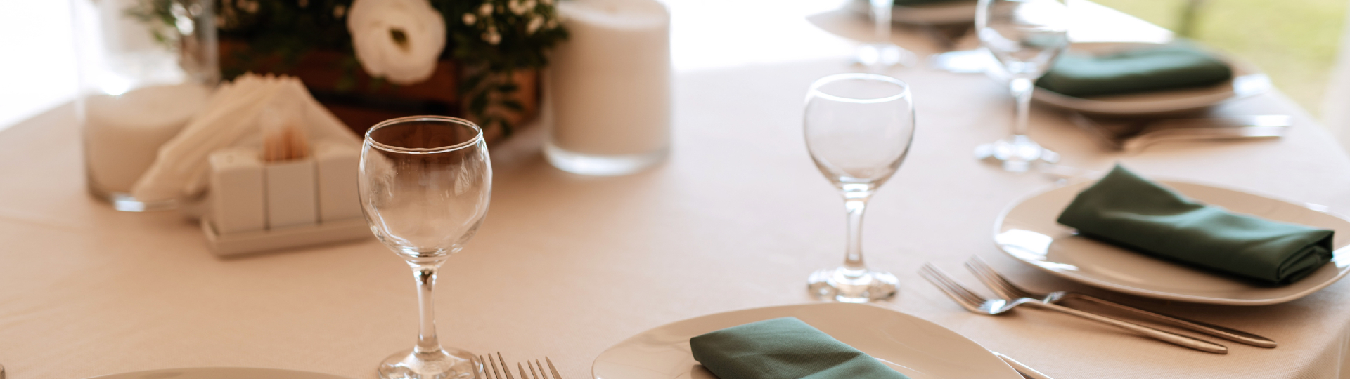 Table set for a formal dinner with white plates, green napkins, wine glasses, and polished silverware on a cream tablecloth.