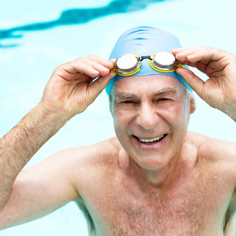 A senior wearing a swim cap and goggles stops swimming to smile for a photo